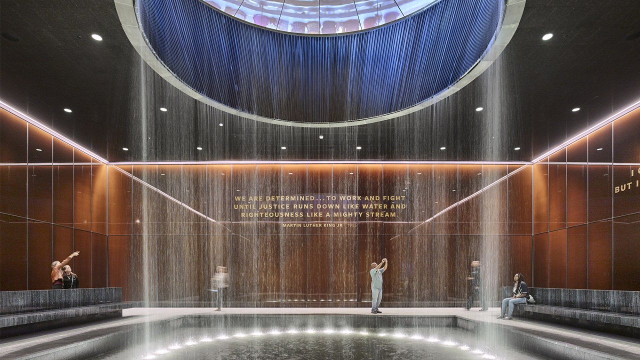 A large, rectangular space is filled with a pool of water into which water falls in a circle from a light-filled skylight in the ceiling. Stone benches line the pool and the back wall bears a quote from Dr. Martin Luther King, Jr. Visitors scattered through the scene point, take photos, and sit in quiet contemplation.