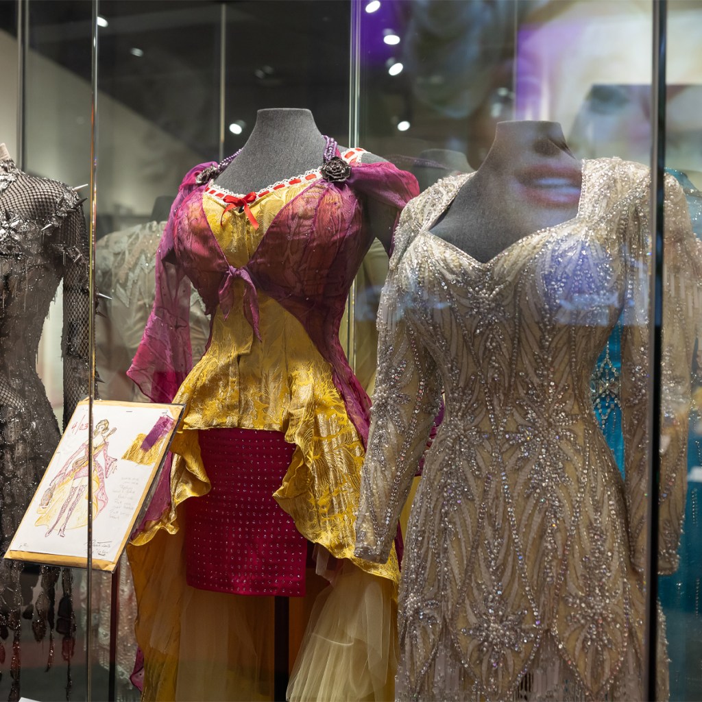 Three sparkly and embellished dresses worn by Dolly Parton displayed in a glass case. On the left is a black and silver metallic dress made of mesh and metal spikes. In the center, is a yellow dress with a pink robe overlaid that ties across the chest. Below it is the designerโs sketch of the dress. The dress to the right is cream and white embellished with silver sparkles.