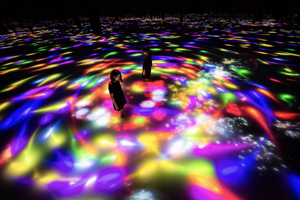 Two women in black dresses stand in an ambiguous black space. Their legs seem to be immersed in water up to their mid-calf. Bright rainbow-colored lights swirl and move across the surface of the water, with a circular eddy around the two figures in the center. 