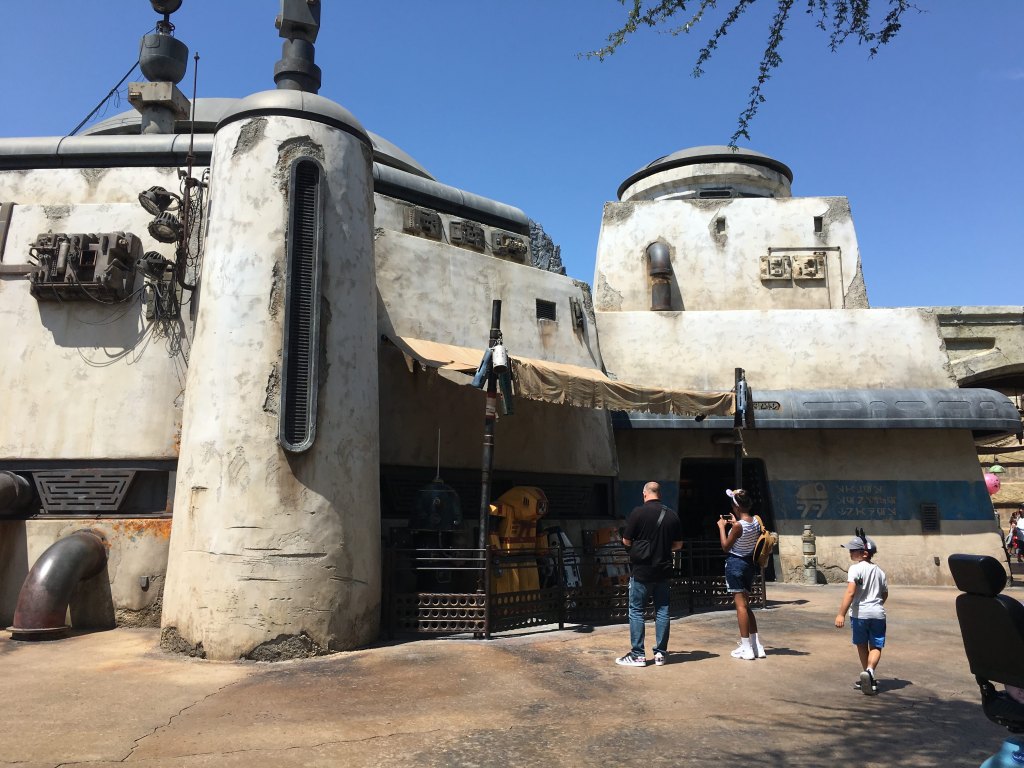Three visitors stand in front of an oddly shaped, white-stucco building beneath a bright blue sky. The building appears worn and weathered, with cracks and stains over its surface. There are no windows and large, black metal pipes, antennae, and other mechanical elements jut from its roof and walls. The three figures are looking at and taking pictures of a row of colorful metal robots that are behind a metal railing in front of the building.