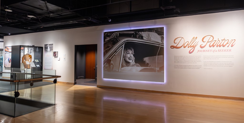 A soft pink light frames a large black-and-white photo of Dolly Parton, a white woman with blonde hair smiling from inside a car. Next to it is the introductory text for the special exhibition, Dolly Parton: Journey of a Seeker.