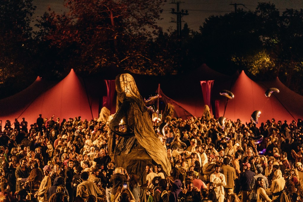 A crowd of people wearing headphones surrounds a large, humanlike puppet made from translucent beige fabric and wire. There are red tent-like structures and trees behind the crowd, which appears to be outside at night.