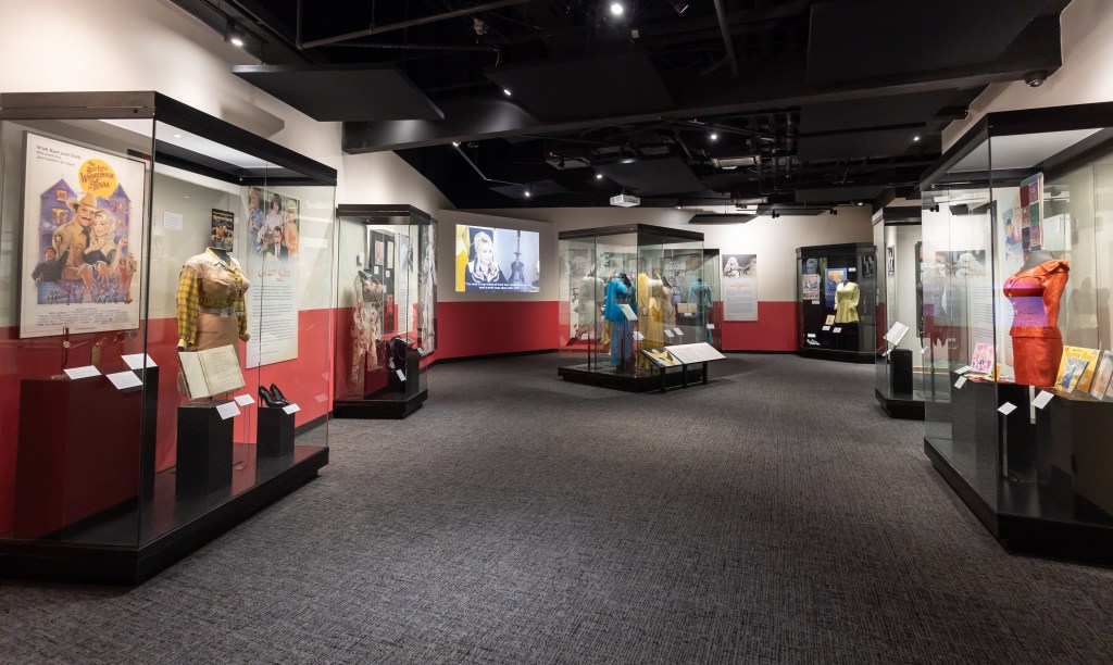 Six large glass-paneled display cases line the walls of an open gallery. Each contains a variety of costumes and collectibles. In the center of the room stands the large glass display case titled โAll Her Colorsโ with six sparkly dresses. A video of a Dolly Parton performance plays on the wall behind it.