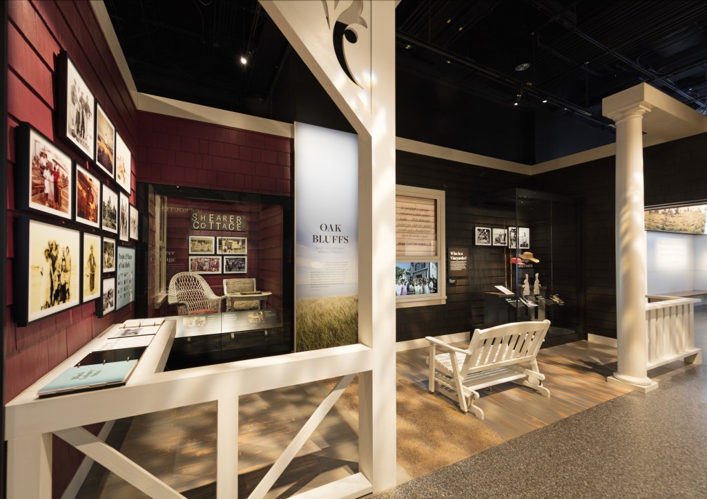 A museum exhibition mimics the look of the front porch of a red-shingled cottage. White columns and a railing section off the exhibition zone, and a white wooden porch rocker occupies the middle of the space, facing a “window” filled with text and an image of a crowd. A text panel to the left of the window reads “Oak Bluffs” with a scene of a beach grass. A display of photographs hangs on the wall to the left with flip books beneath and a display of wicker furniture bears the sign (Shearer Cottage). Additional artifacts are displayed in a case to the right. 