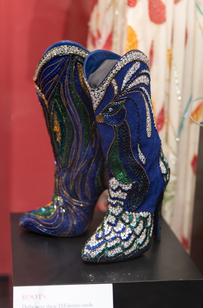 A pair of mid-calf high-heeled boots made of blue suede. They are embellished with sparkly beads forming a green, purple, white, and blue peacock. The peacockโs head and neck is on the left boot while its tail is on the right boot.