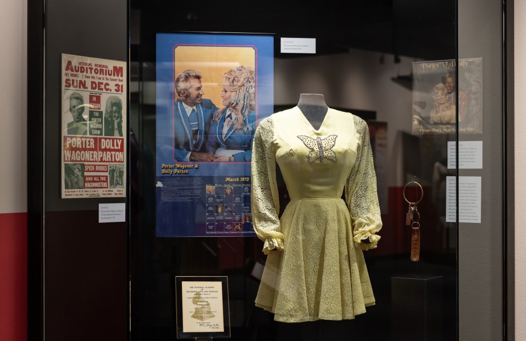 A glass display case features a long-sleeved yellow dress styled onto a black, headless mannequin. The front of the dress has a silver sequin butterfly. To the left of the dress are two posters. One is white, green, and red advertising a performance of The Porter Wagoner Show on December 31 at Veterans Memorial Auditorium. The other poster is blue and features a young Dolly Parton in a white shirt and blue suit jacket with blue ribbons in her hair. She is facing a white man with blonde hair wearing a similar blue and white jacket. To the right of the dress is a record album with the same man and woman on it titled Porter โnโ Dolly. Below that is a large gold ring with keys and a brown leather strap embossed with โDolly.โ
