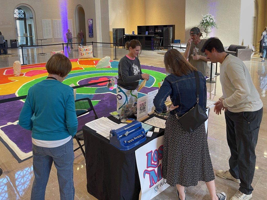 Visitors stand beside and interact with a large, brightly colored labyrinth-style installation in a museum gallery, engaging with the work through movement, observation, and conversation.