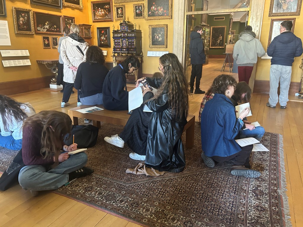 A group of young adults sits on a colourful rug on the floor of a museum gallery. They are all holding sketch pads and drawing. On the walls are paintings of colorful flowers, insects, and fruit in bright colors.