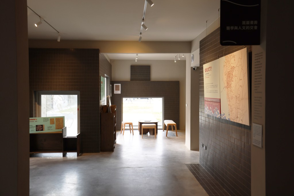 An empty renovated residence interior with display cases, wall panels, track lighting, and a small seating area near a window. A brown hanging sign reads “Kun-Yen Study” beside the plague exhibition area.