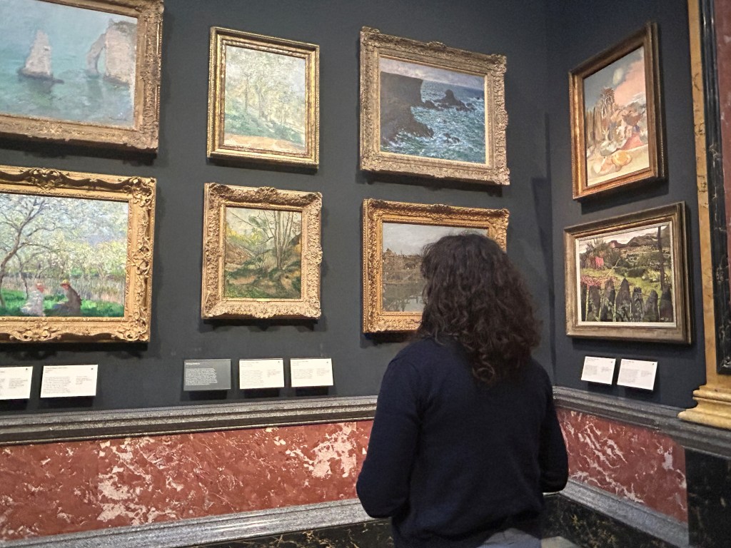 A young woman with curly dark hair stands with her back to the viewer concentrating on a wall of landscape paintings in a museum gallery. The paintings show woods, beaches, and orchards; each one is in an ornate gold frame on a dark-gray wall.