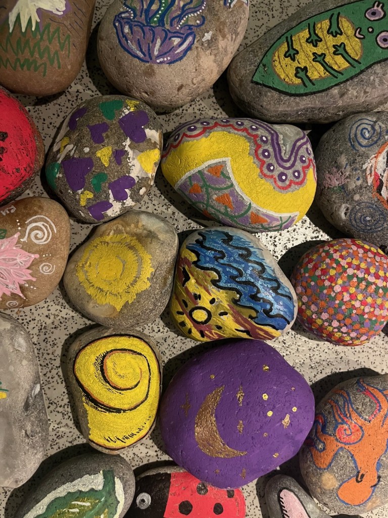 A close-up image of colorful, smooth rocks painted with various images including a snail, a beach scene, a jellyfish, and an octopus.