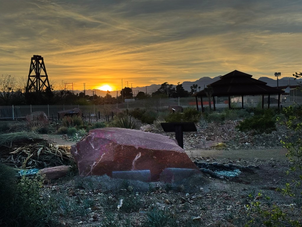 At dusk, a large red stone boulder in the foreground is surrounded by plants. A ramada and well derrick in a garden can be seen with the sun setting behind mountains in the background.