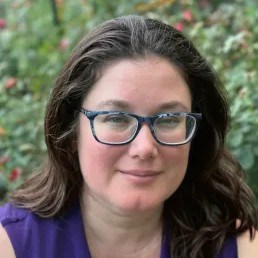 A woman with shoulder length brown hair and glasses looks at the camera.