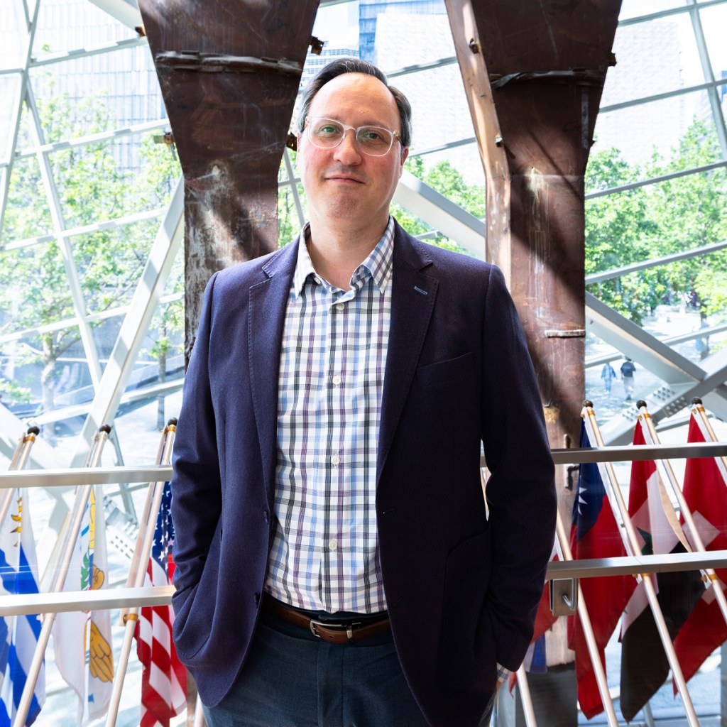 A fair-skinned man wearing wire-rim glasses, a navy-blue blazer and pants, and a checked shirt stands with his hands in his pockets facing the camera in front of a display of in a glass-enclosed museum atrium.