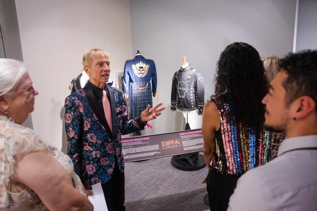 An older white gentleman faces the camera in a black shirt, pink tie, and custom-embroidered and beaded jacket talking to three visitors. In the background are three mannequins with jackets from his collection that have custom artistic designs on them, including two with skull motifs.Fig. 2. Community partner and lender Mike Matthews speaks with visitors about his collection of DIFFA (Design Industries Foundation Fighting AIDS) jackets that are created to raise funds for HIV/AIDS care and prevention. 