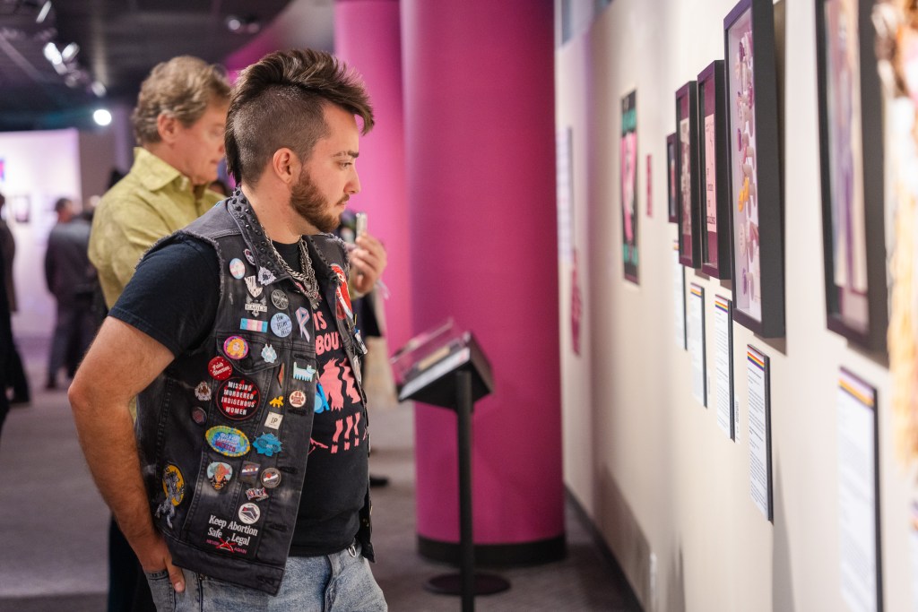 Visitors face a wall of framed artifacts on view in the exhibition. The visitor in the foreground has a brunette mohawk and wears a black denim vest that is artfully decorated in various pinback and enamel buttons. Bright pink architectural columns are visible in the background.
