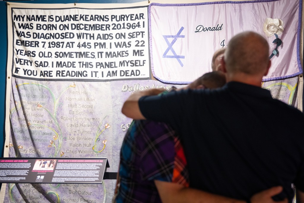Visitors embrace with their backs to the camera while viewing panels from the National AIDS Memorial Quilt, including one in the upper left that reads in all-capital letters, in a bold black font sewn onto a white background, “My name is Duane Kearns Puryear. I was born on December 20, 1964. I was diagnosed with AIDS on September 7, 1987 at 4:45 pm. I was 22 years old. Sometimes it makes me very sad. I made this panel myself. If you are reading it, I am dead.”