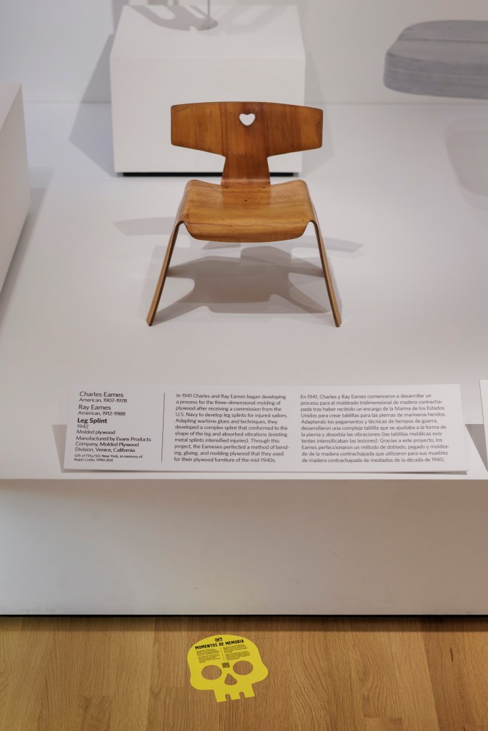 A small, molded plywood chair, titled **Childโs Chair** (about 1944), with a heart-shaped hole in its backrest by Charles and Ray Eames sits on a white platform with a bilingual label in front of it; in the foreground on the wooden floor lies a yellow, two-dimensional paper skull.