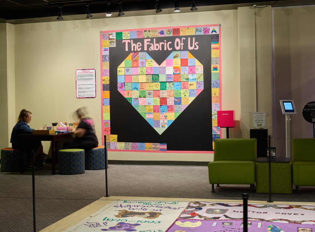 Image of exhibition gallery. In the foreground, a section of the National AIDS Memorial Quilt is shown on a platform, surrounded by stanchions. The focal point of the photograph is a multicolored installation on the wall titled “The Fabric of Us.” The installation has a black background with square colored pieces of paper assembled in the shape of a heart. Two visitors are seated at a table to the left creating their own submissions for the wall.
