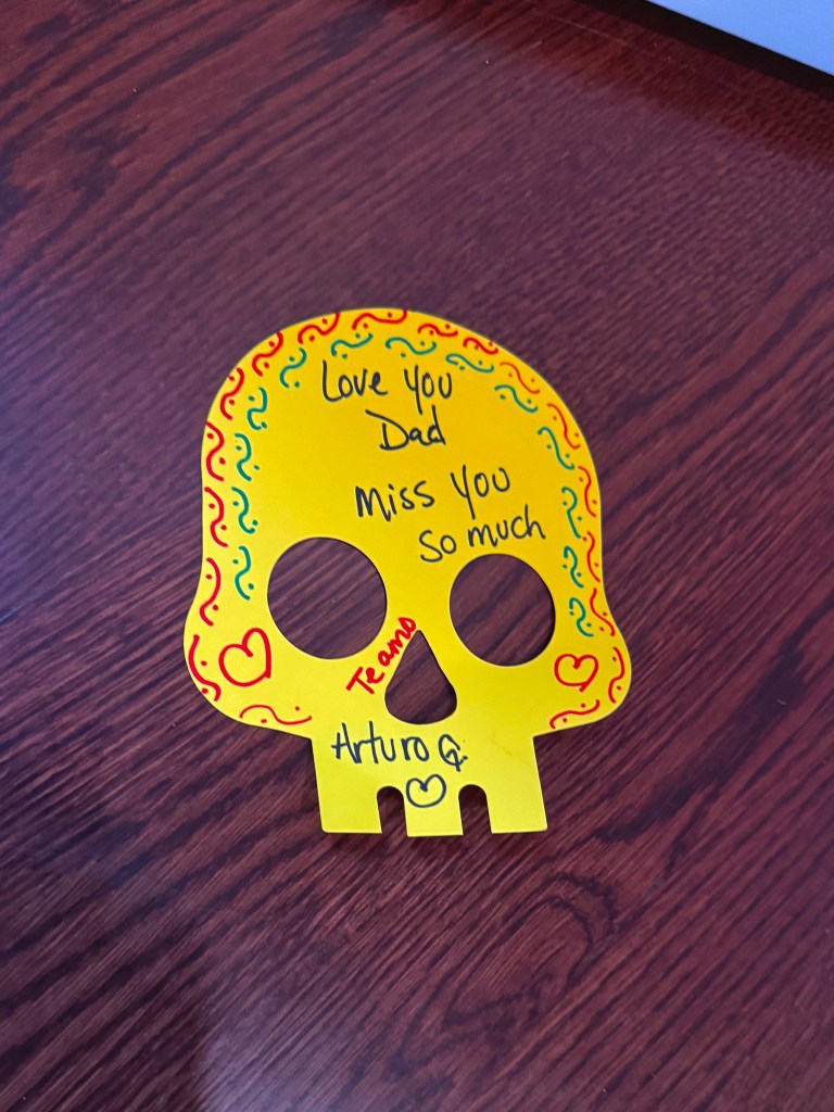 A yellow, two-dimensional paper skull rests on a dark wood floor. The skull is decorated by a visitor with hand-drawn red and green swirls and dots along the border, one red heart on each cheek, and a black heart on the bottom of the skull. The visitor also wrote on the skull. In black marker, the text reads, โLove You Dad / Miss You So Muchโ on the forehead of the skull. Near the nose of the skull, in red marker, the text reads, โTe amo.โ At the bottom of the skull, in black marker just above the black heart, the text reads, โArturo G.โ