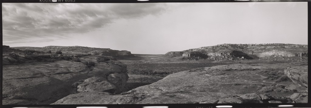 A black-and-white panoramic photograph depicting a wide, sweeping view of Chaco Canyon from the north mesa, looking south through a gap in the canyon walls. Rocky, textured stone formations fill the foreground. The canyon floor, filled with vegetation, stretches into the middle distance, flanked by mesa cliffs on either side, beneath an open sky with clouds.