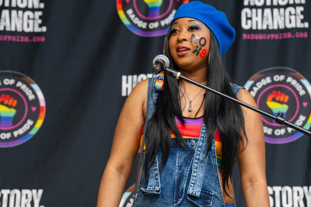 Mz. Trill, an African American spoken word artist, stands at a microphone and delivers her work “No H8.” She has a temporary tattoo on her cheek with the letters “NO H8.”