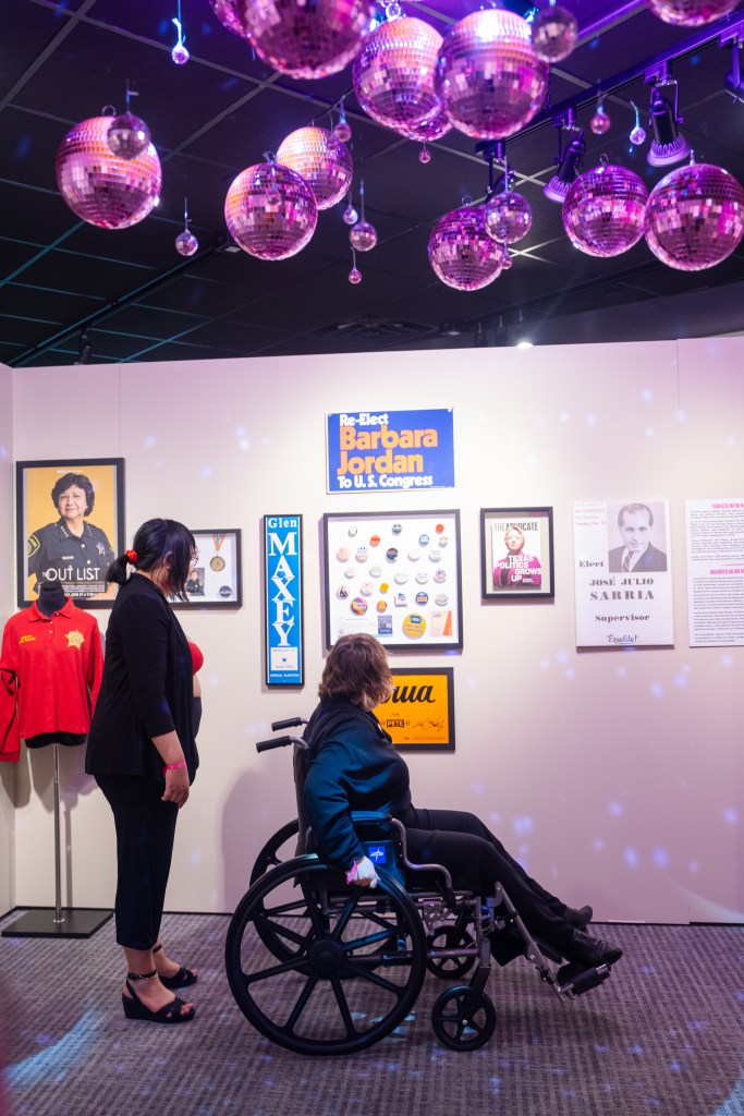 Two visitors, one in a wheelchair, view a section of the exhibition that highlights political campaign materials including a poster of Dallas County Sheriff Lupe Valdez, campaign posters for Barbara Jordan and Glen Maxey, and a shadowbox of pinback campaign buttons. Pink disco balls can be seen hanging from the ceiling.