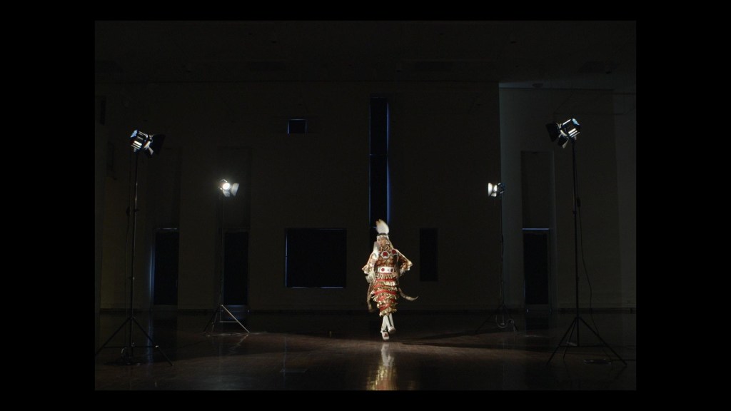A dark, dramatically lit video still showing a woman dancing alone in a large empty interior space. The dancer wears red, white, and gold jingle dress regalia with a white and red beaded belt, white beaded leggings, and white feather plume. Her left hand is placed on her waist, and she holds a feather fan in her right hand. Four studio lights flank her, casting light onto her and a dark, reflective wood floor. The surrounding space is largely in shadow.